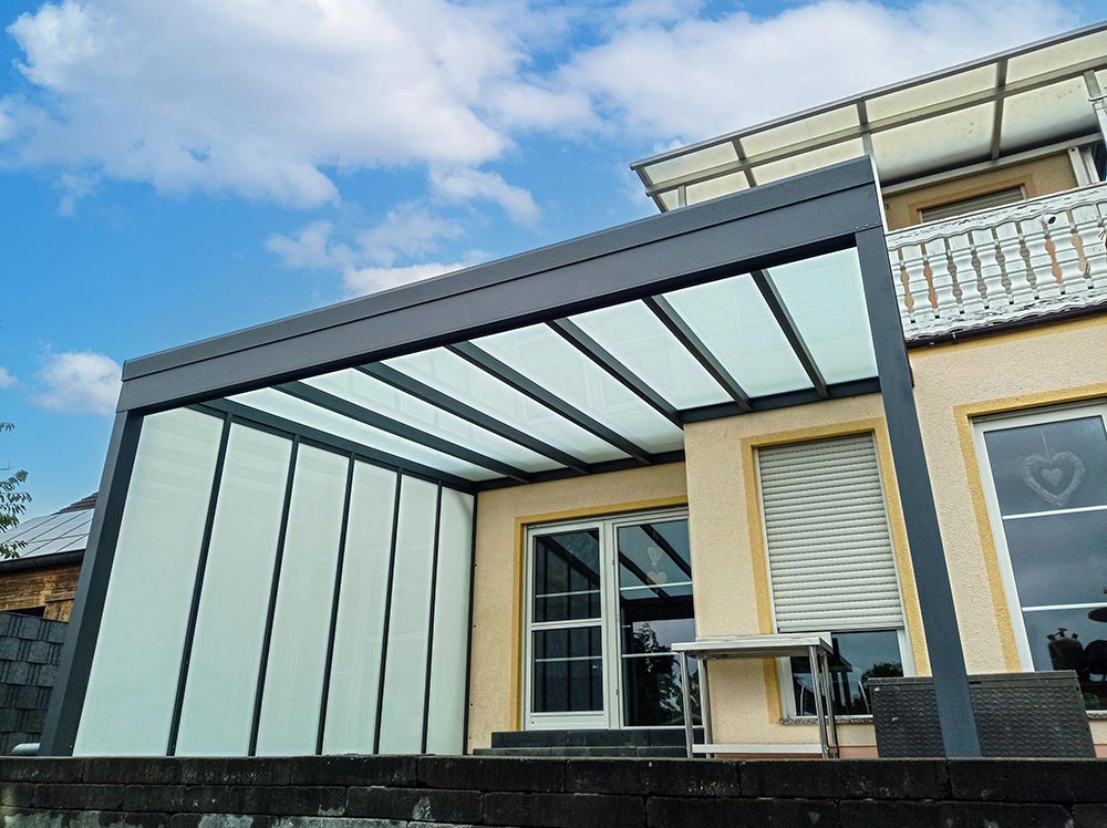 Modernes Terrassendach mit Glas und grauen Streben an einem gelben Haus bei Sonnenschein