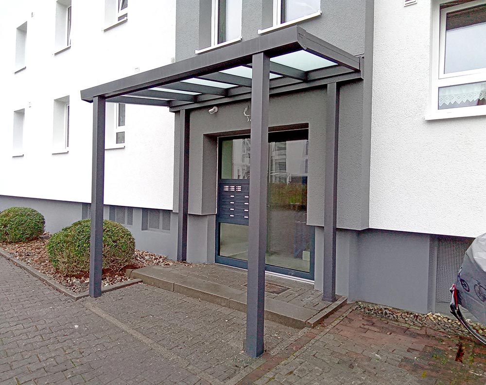 Moderner Eingang mit grauem Glasvordach, weißen Wänden und dunkler Haustür